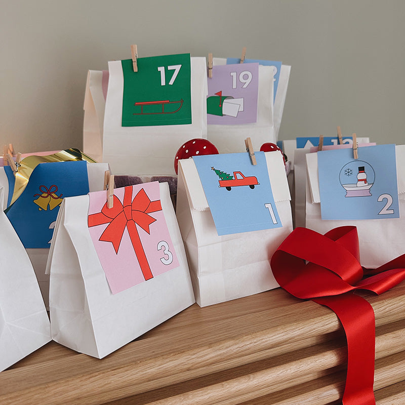 Selbstgemachter Adventskalender aus weißen Papiertüten mit bunten Zahlenkarten und großer roter Schleife – DIY-Vorlage von Gutschein-Werkstatt.
