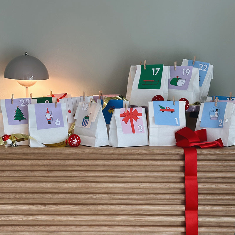 DIY-Adventskalender mit weißen Papiertüten und farbigen Anhängern, dekoriert mit rotem Geschenkband – moderne Bastelidee von Gutschein-Werkstatt.